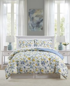 a white bed with yellow and blue flowers on it in front of two windows next to nightstands