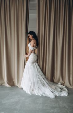 a woman standing in front of a curtain wearing a wedding dress with an off the shoulder neckline