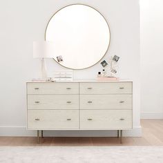 a white dresser with a round mirror above it and a lamp on the sideboard