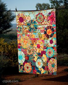 a colorful quilt hanging on a clothes line in the sun with trees and bushes behind it