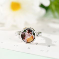 This photo ring is the best choice to make the memory eternal. Fashioned in sleek sterling silver, this ring features a charming daisy, and a shimmering round-cut stone is set at the center to complement the brilliance. With your own photo included, this beautiful piece will be more memorable. You can also wear it as your pendant to make your daisy-themed necklace dangling close to your heart.Carat Weight: 0.81 ctStone Size: 3,1.5,1.3 mmStone Type: Jeulia® StoneNumber of Stones: 28 Stone Shape: RoundStone Color: Citrine Yellow, Diamond WhiteWeight: 4.6 gWidth: 11.4 mmHeight: 5.9 mmThickness: 3.1 mmMaterial: 925 SilverPlating Color: Silver, Rose Gold Classic Round Birth Flower Jewelry, White Gold Jewelry With Rotating Bezel As Gift, Elegant White Gold Round Pendant Ring, Sterling Silver Rings With Rotating Bezel For Anniversary, Silver Rings For Mother's Day, Dainty Birth Flower Ring, Fine Jewelry Ring With Rotating Bezel, Mother's Day Sterling Silver Engraved Ring, Dainty Flower Ring With Birthstone