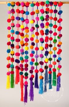 a multicolored wall hanging with tassels and beads on the bottom, in front of a pink background