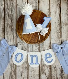 "Celebrate your little one's first birthday with this classic blue seersucker party decor set! So cute for cake smashes and photos.  \"ONE\" banner is perfect for a high chair, wall display, or fireplace mantel.  Length from end to end is approximately 60\". Blue seersucker letters are appliquéd and embroidered on white fabric. Party hat coordinates with the banner. Elastic strap secures the hat on baby's head either under their chin or behind their head like a headband. Cake topper is blue and white seersucker with an array of white ribbons at the bottom. Comes with a clear plastic stick to push into the cake.  Please feel free to message me with any questions!" Blue And White 1st Birthday Theme, Boys 1st Birthday, Outdoor First Birthday Party, Blue And White First Birthday Cake, One Is Fun Birthday, Classic Blue First Birthday, Classic Blue And White First Birthday, Blue Gingham First Birthday, Light Blue Smash Cake