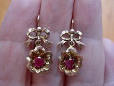 "These antique earrings are hard to date. The bow motif was popular in the Georgian era and again in the late Victorian/Edwardian era. I am guessing the latter. They have bow shaped tops and a flower dangle base. Each flower has a .30 carat natural ruby. These earrings are small and measure about 1\" from the wire tops. One earring is missing its small wire catch. The piece is there and can be soldered back into place or wear them as is. I will include a plastic stopper to use so not to lose the Yellow Gold Bow Earrings For Wedding, Classic Bow Earrings For Anniversary, Vintage Wedding Jewelry With Bow Detail, Victorian Screw Back Earrings For Wedding, Victorian Screw Back Earrings For Formal Occasions, Victorian Screw Back Wedding Earrings, Victorian Wedding Earrings With Screw Back, Victorian 14k Gold Earrings For Anniversary, Victorian 14k Gold Earrings