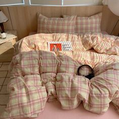 an unmade bed with pink and yellow plaid comforter on the floor next to a lamp