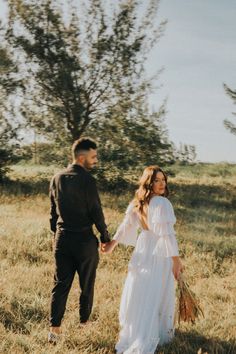 a man and woman holding hands while walking through a field with trees in the background