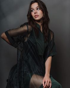 a woman sitting on top of a chair in a green shirt and black pants with her hands behind her head