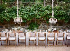 an outdoor dining table with chandeliers hanging from the ceiling and greenery behind it