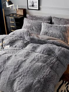 a bed covered in grey fluffy blankets and pillows with a lamp on the nightstand next to it