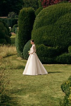 a woman in a wedding dress is walking through the grass by some bushes and trees