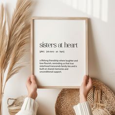 two hands holding up a framed print with the words sisters at heart on it, next to some straws