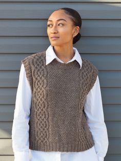 a woman standing in front of a gray wall wearing a brown vest and white shirt