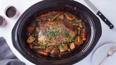 a slow cooker with meat and vegetables in it on a white tablecloth next to utensils