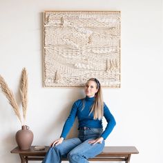 a woman sitting on top of a wooden bench next to a vase and wall hanging