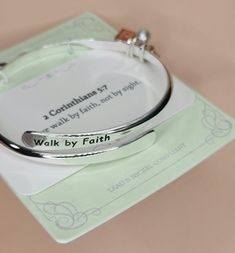 This silver bangle bracelet wraps around to overlap and there you'll find the imprint of "Walk by Faith" in black. Dangling from the bracelet are 3 charms - a gold cross, a pearl and a copper Bible. Silver Bangle Bracelet, Walk By Faith, Silver Bangle Bracelets, Silver Bangle, Gold Cross, Walking By, Silver Bangles, Bangle Bracelet, Bangle Bracelets