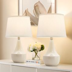 two white vases sitting on top of a table next to a lamp and a book