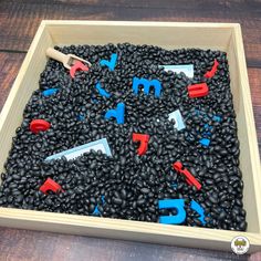 a wooden box filled with lots of black beans and plastic letters that spell out the word m