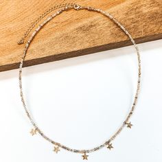 This is a beaded necklace with gold bead spacerss and five gold star charms. This neckalce includes a nude mix of beads. This necklace is pictured laying partially on a wood block on a white background. Gold Star Earrings, Giddy Up Glamour, Layering Pieces, Star Charms, Gold Stars, Star Earrings, Delicate Bracelet, Beaded Necklace, Stars