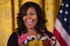 the first lady smiles while standing in front of an american flag and yellow curtained background