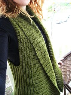 a woman wearing a green knitted vest and black shirt standing in front of a chair