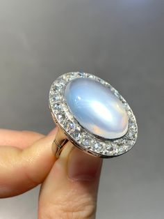 "A dreamy glowing moonstone cabochon ring measuring 2.7 cm by 2.1 cm, encrusted with approximately 1.8 carats of glimmering chunky icy-white rose cut diamonds. The moonstone shines out a eye-catching luminous blue adularescense. Currently measures a size 6. Ring Face Measurement: 2.7 cm by 2.1 cm Measurement of Moonstone: 2.0 by 1.1 cm Approximate Carat Weight of Diamond: 1.8 Carats Gold Purity: 14K Markings: \"14K\" Ring Size: 6.75 (Resizable)" Faceted Moonstone Ring, Oval Diamond White Moonstone Ring, Oval Moonstone Ring With Diamond Halo Setting, Heirloom Diamond Moonstone Ring In Oval Shape, Heirloom Diamond Oval Moonstone Ring, Heirloom Oval Diamond Moonstone Ring, Heirloom Oval Moonstone Diamond Ring, Fine Jewelry Oval Moonstone Ring With Rose Cut Diamonds, White Oval Moonstone Ring With Rose Cut Diamonds