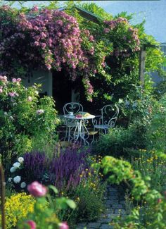 an outdoor table and chairs in the middle of a garden with flowers on either side