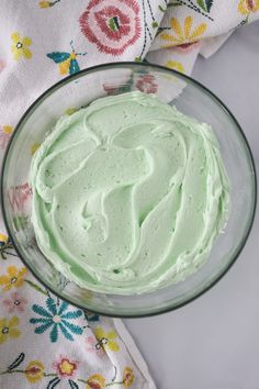 a glass bowl filled with green frosting on top of a floral table cloth next to a napkin