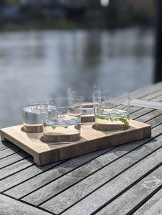 four glasses on a wooden tray with water and plants in the middle, near a body of water