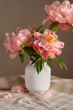 three pink flowers in a white vase on a plaid tablecloth with petals scattered around