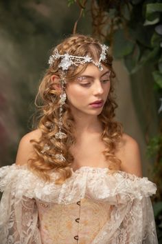 a woman with long curly hair wearing a white dress and headpiece in front of greenery