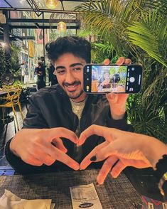 a man sitting at a table with his hands in the shape of a heart while taking a selfie