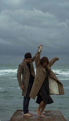 a man and woman standing on top of a rock next to the ocean with their arms in the air