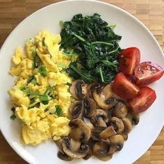 a white plate topped with scrambled eggs, mushrooms and tomatoes next to spinach on a wooden table