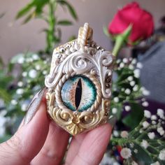 a hand holding a miniature eye in front of a vase with flowers and greenery