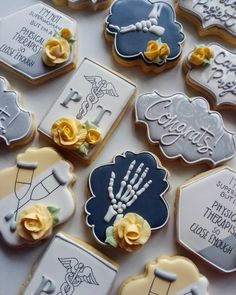 many decorated cookies are arranged on a table with white and blue icing, yellow roses, and handwrittens