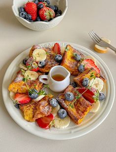 a white plate topped with french toast and fruit next to a bowl of strawberries