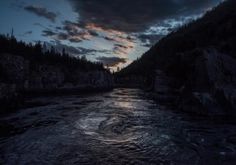 the sun is setting over a river with rocks and trees in the foreground, while dark clouds loom overhead