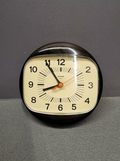 a black and white clock sitting on top of a gray floor next to a wall