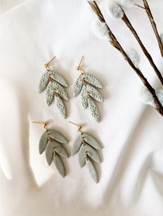 three pairs of green leaf shaped earrings on top of a white cloth next to branches