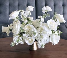 white flowers in a gold vase on a wooden table next to a gray velvet couch