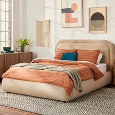 a bed with an orange blanket and pillows in a white room next to a brick wall