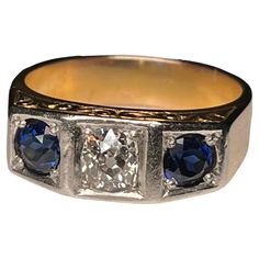 This is an Art Deco 14K Yellow Gold, Diamond and Sapphire Garter Ring. It depicts a gold ring with a top divided by two small squares at the sides and one larger one in the center. The center one is embellished by an old European brilliant diamond ( Weight- .75 carats; Measurements- 5.10 x 5.0 x 2.94 mm; Color- G; Clarity- SI-2; Type- Kimberly) and the side ones by two sapphires each one is weighing 0.5 carats. The ring is also decorated with an engraved branch of scrolls at the front and back o 14k Yellow Gold Square Cut Diamond Ring, Heirloom Rectangular Brilliant Cut Rings, Formal Gold Three Stone Rings, Art Deco Yellow Gold Three-stone Ring, 14k Gold Rectangular Brilliant Cut Jewelry, 14k Gold Rectangular Jewelry With Brilliant Cut, Heirloom Rectangular Brilliant Cut Diamond Ring, Rectangular Brilliant Cut Sapphire Ring For Formal Occasions, 14k Gold Jewelry With Brilliant Cut Rectangular Shape