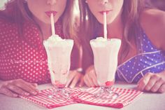 two women sitting at a table with drinks in front of their faces, one drinking milkshakes