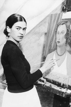 a black and white photo of a woman in front of a painting holding a paintbrush