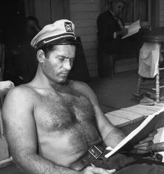 a shirtless man sitting on a bench reading a book while wearing a sailor's hat