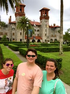 three people standing in front of a large building with trees and bushes on both sides