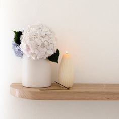 a white vase filled with flowers sitting on top of a wooden shelf next to a lit candle