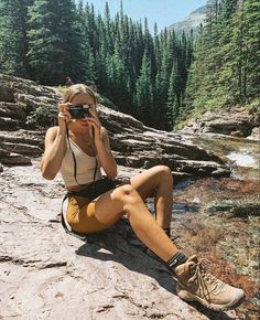 a woman sitting on top of a rock while holding a camera up to her face