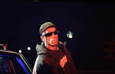 a man wearing sunglasses and a hoodie standing next to a parked car at night