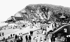 an old black and white photo of people walking on the beach in front of a mountain